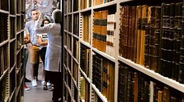 Foto de Estante com vários livros e ao fundo alguns estudantes conversando.