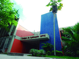 Imagem mostra fachada do Colégio de Aplicação da UFPE. Na imagem, o centro é mostrado junto às árvores e plantas que ornamentam sua fachada nas cores vermelho e azul.